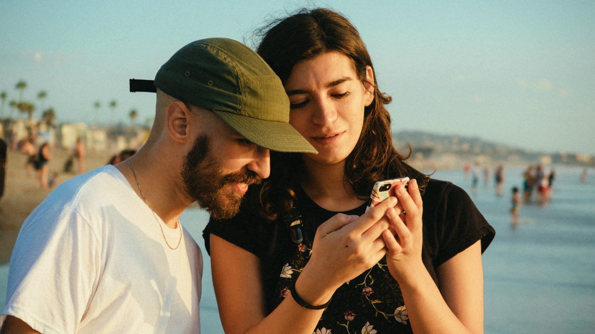 Two people standing close together on a beach at the end of the day, both looking down at one phone that one of them is holding