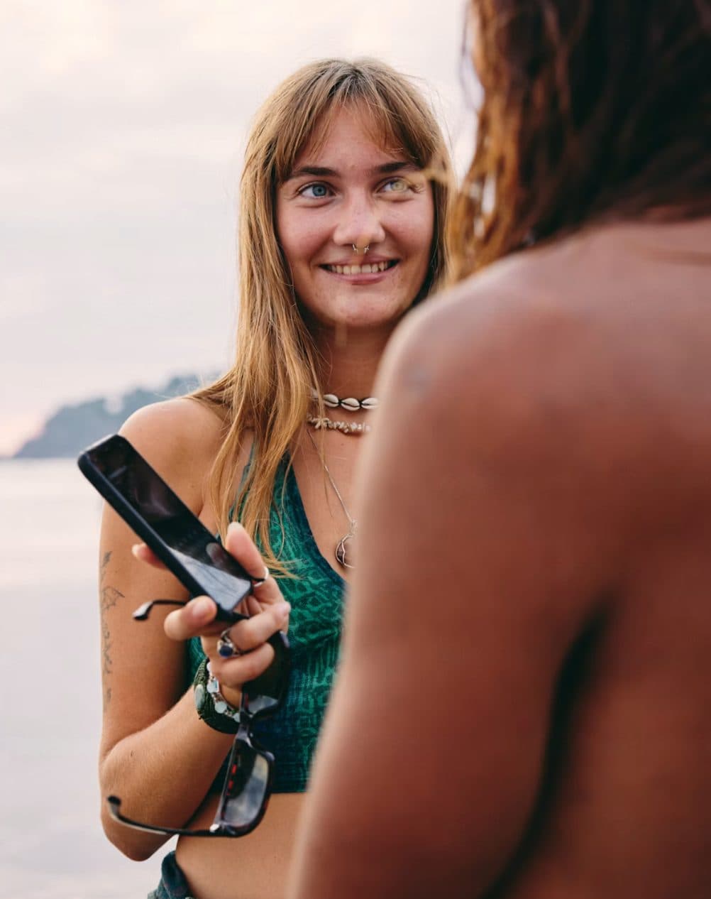 A woman by the sea holding out a phone towards someone standing in front of her, who is turned away from the camera