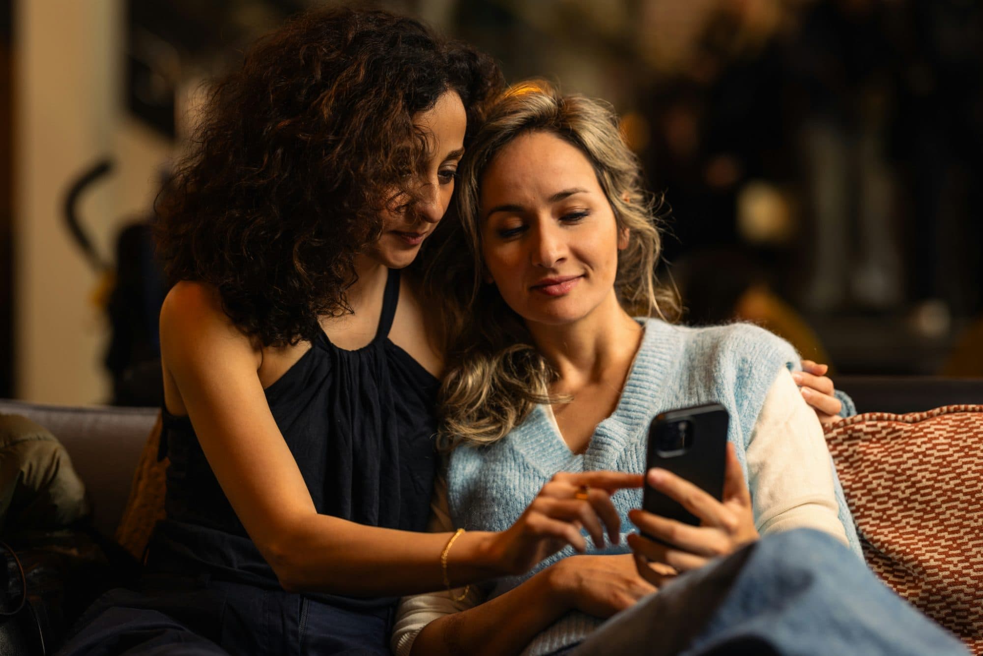 Two women sitting close on a sofa in a warmly lit room, leaning in to look at a phone one of them is holding