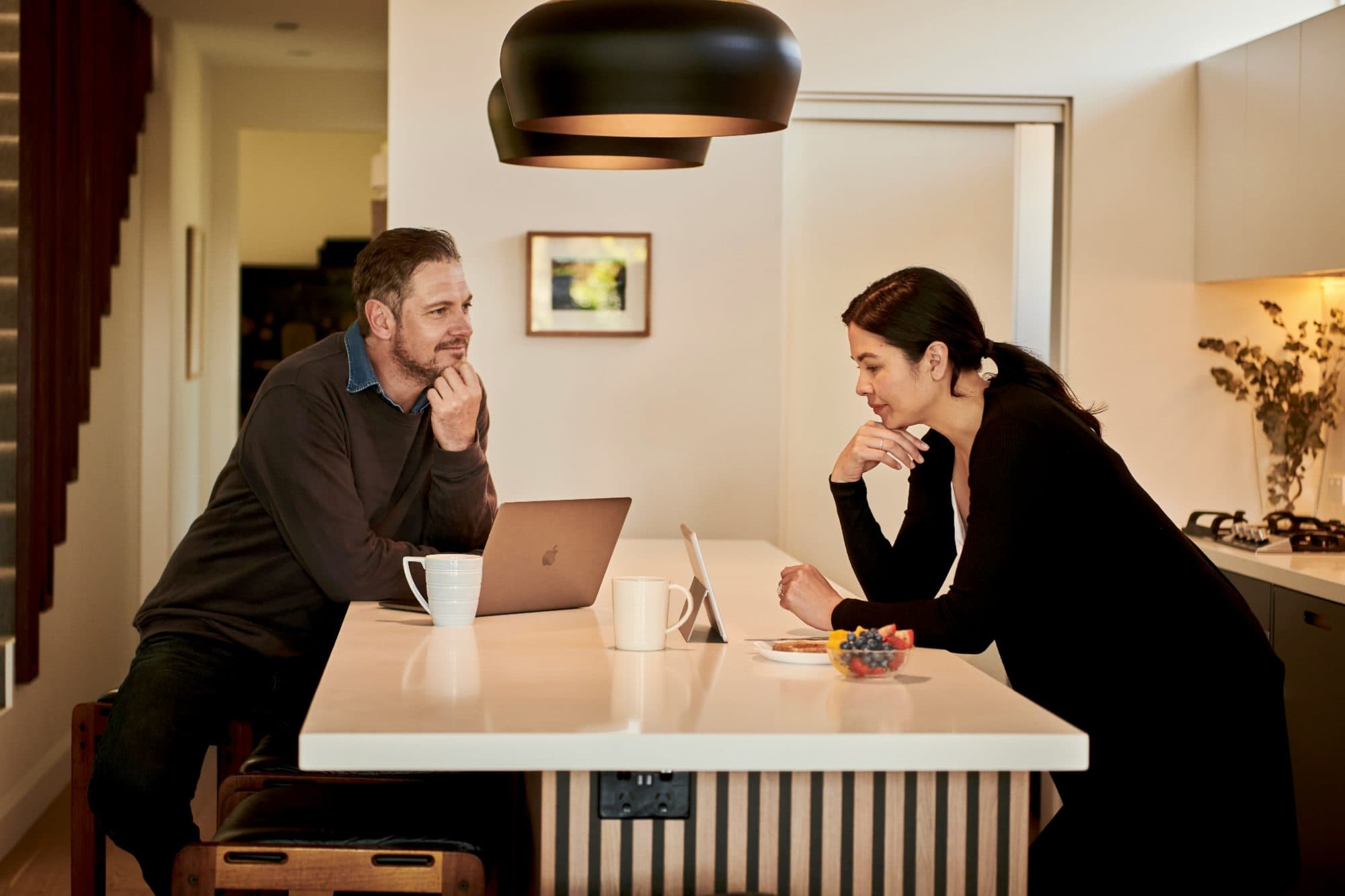 Two people leaning on opposite sides of a kitchen island, talking, with coffee mugs and a closed laptop between them
