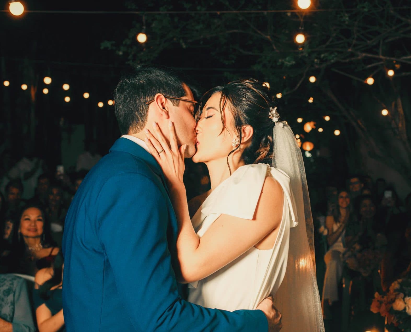 A couple kissing at their wedding ceremony in the evening, under strings of warm lights, with guests behind them