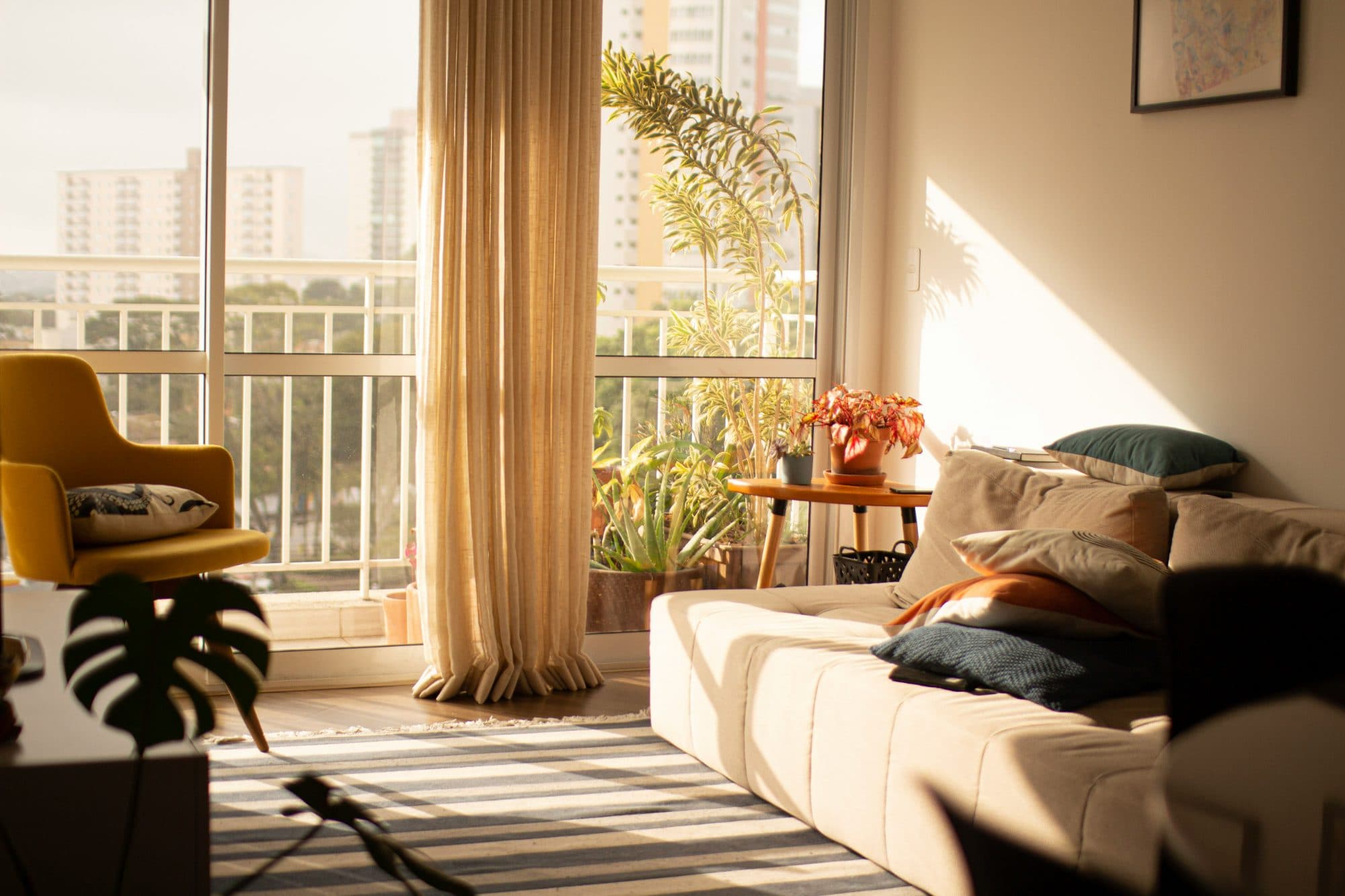 An empty living room in late afternoon light, with a sofa and cushions, a yellow armchair, a striped rug and potted plants on a balcony beyond the open windows