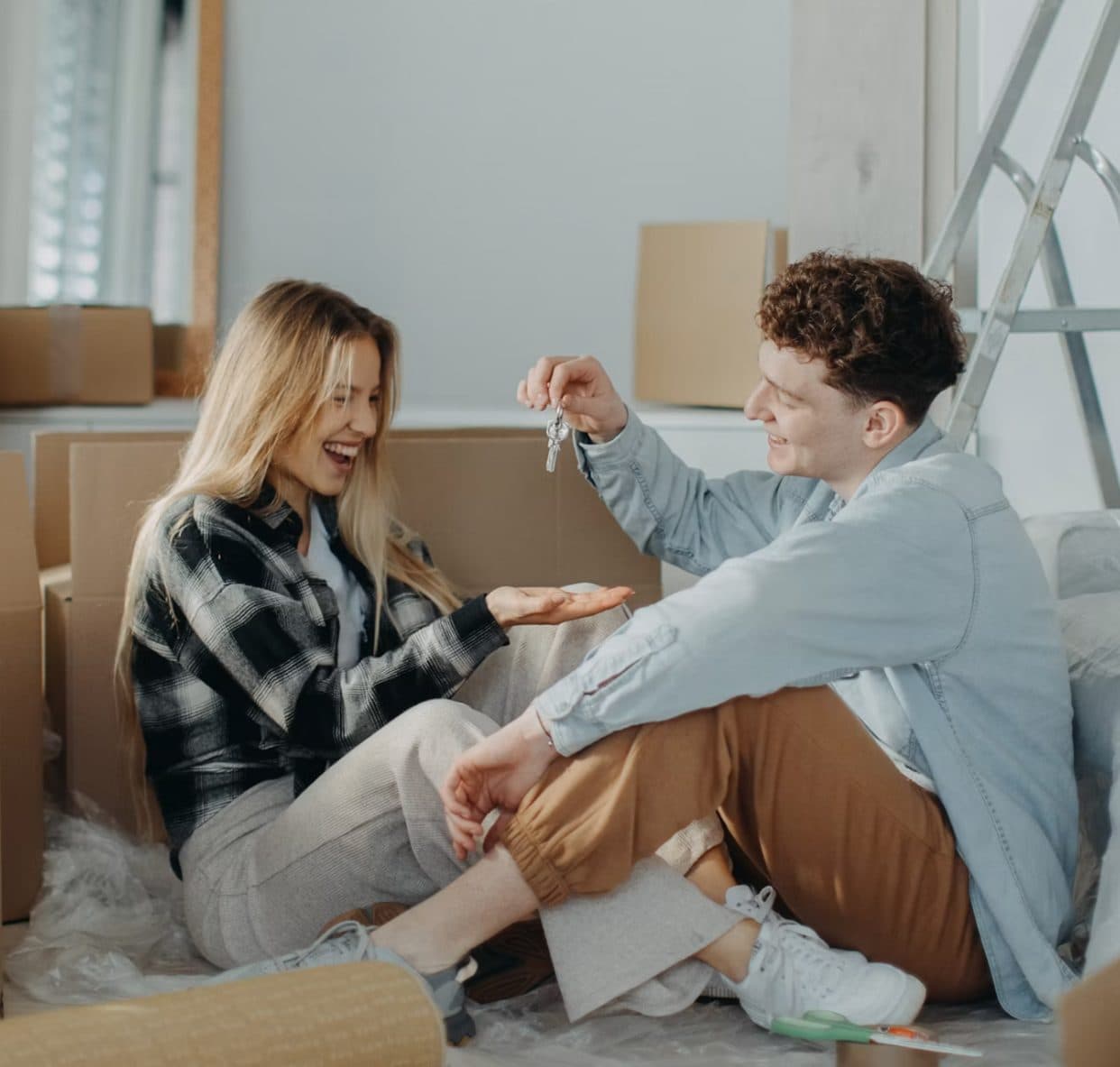 A couple sitting on the floor of a bare room surrounded by cardboard boxes and a stepladder, one passing a set of keys to the other