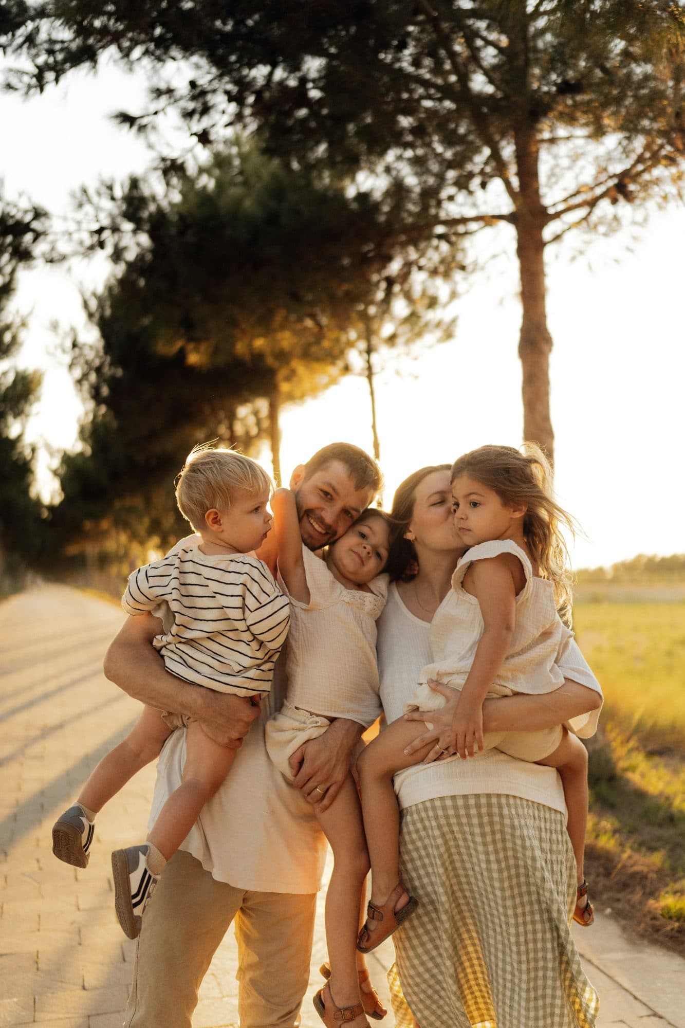 Two parents outdoors at sunset, each carrying a small child, with a third leaning against them