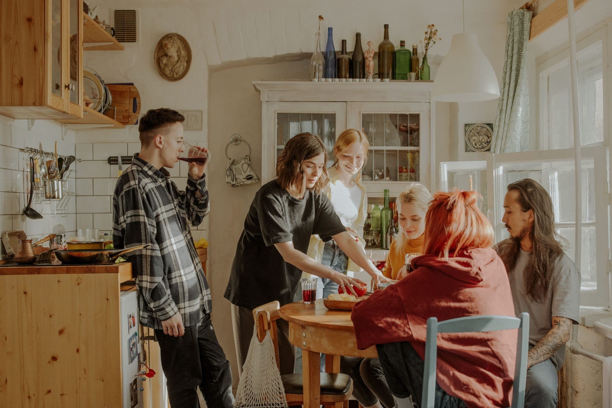 Six people in a small lived-in kitchen in afternoon light, some standing, some at a round table, one pouring a drink