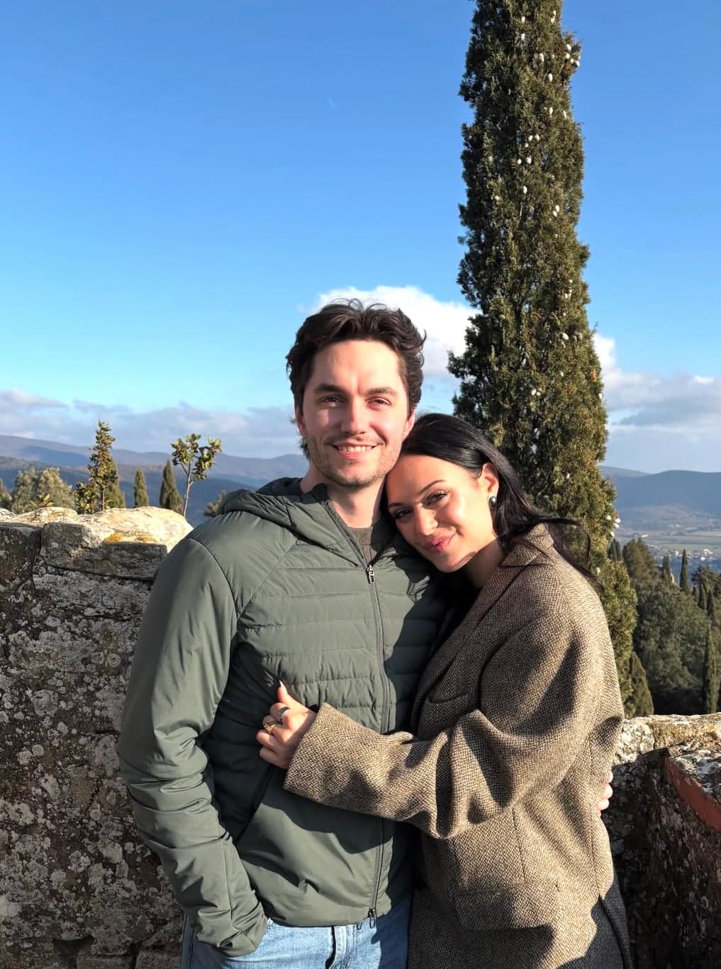 Jason and Lily standing close together by a low stone wall on a hillside in the California backcountry, with cypress trees and rolling hills behind them